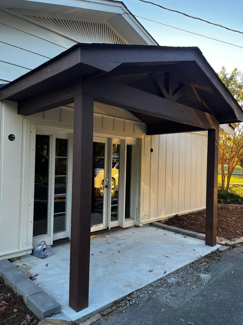 Modern home entrance with a dark wooden canopy, framed glass doors, and newly poured concrete.