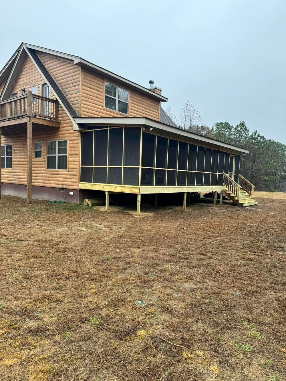 Wooden house with a large screened porch, surrounded by grass and trees under a cloudy sky.