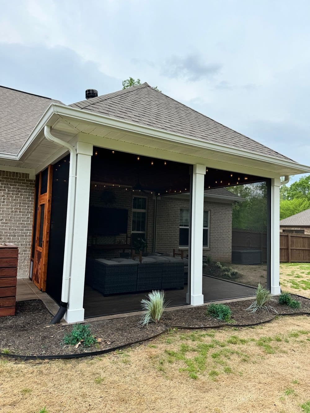 Outdoor screened-in porch with cozy seating and string lights surrounded by landscaped yard.