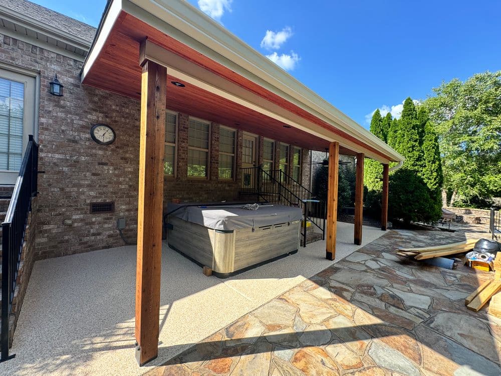 Backyard patio with hot tub under wooden awning, stone floor, and landscaped greenery.