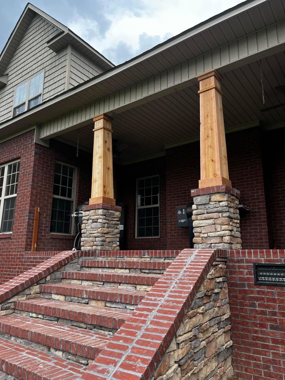Front porch of a brick house with stone columns and wooden beams, featuring brick steps.
