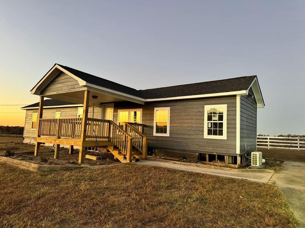 Gray single-story house with a wooden porch and sunset reflections on windows.