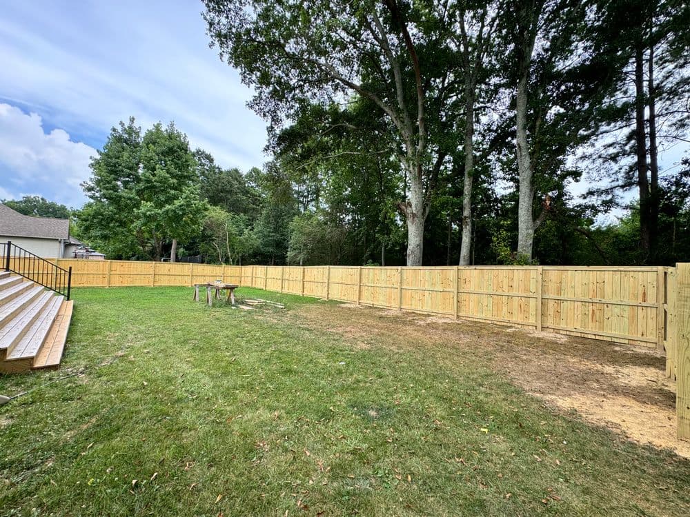 Wooden fence in backyard with trees, patio steps, and grassy area under a blue sky.
