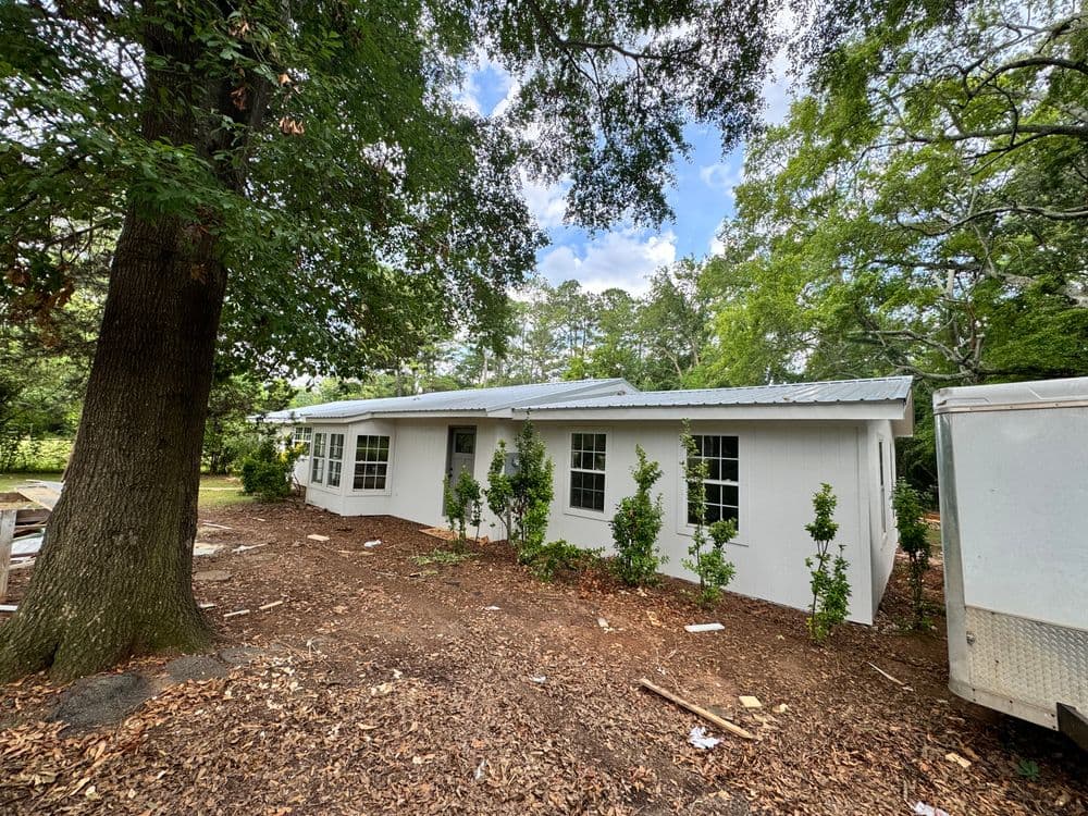 White single-story house surrounded by greenery and mulch under a cloudy sky.