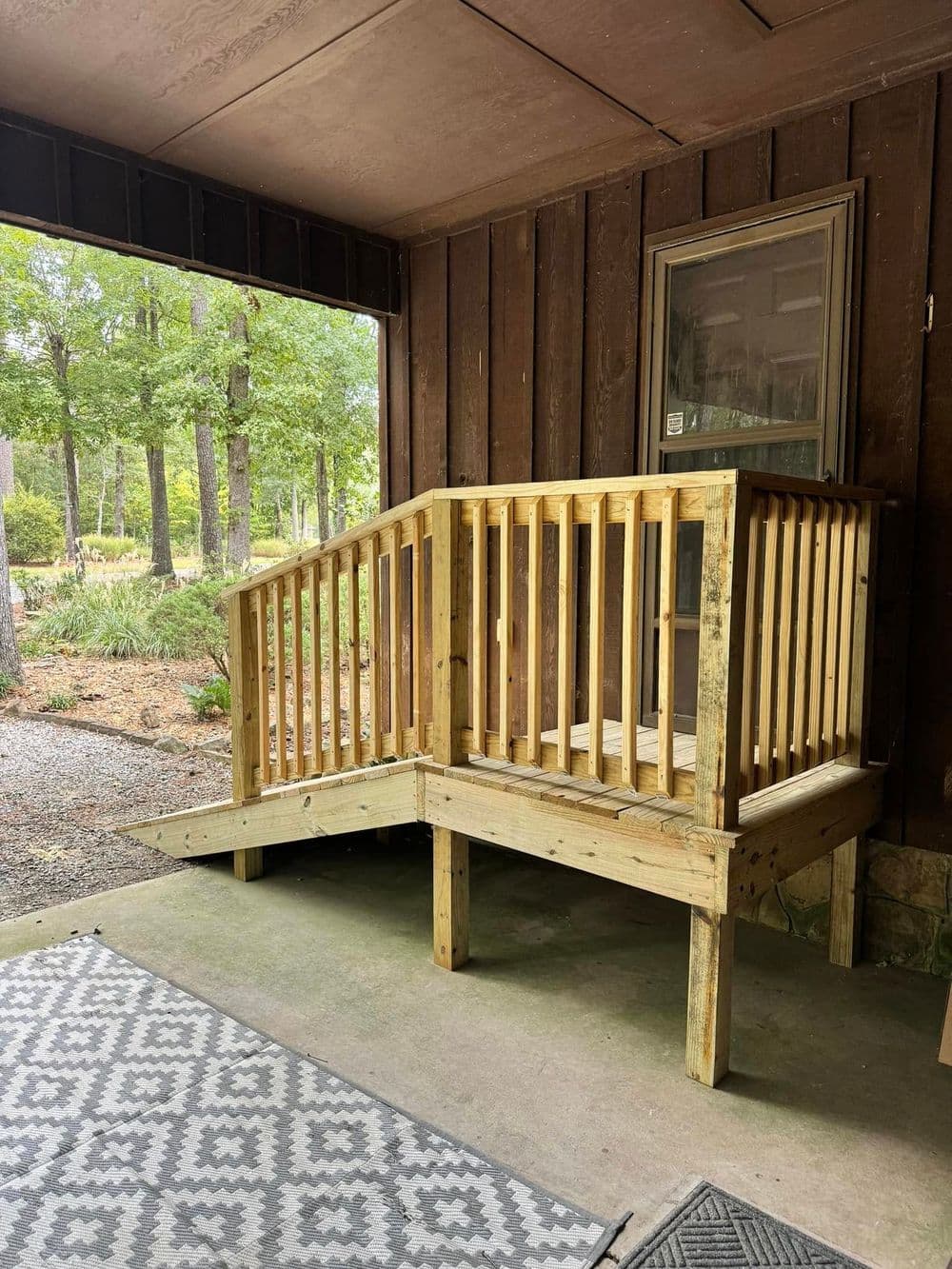 Wooden porch ramp with railing for accessibility, set against a wooded background.