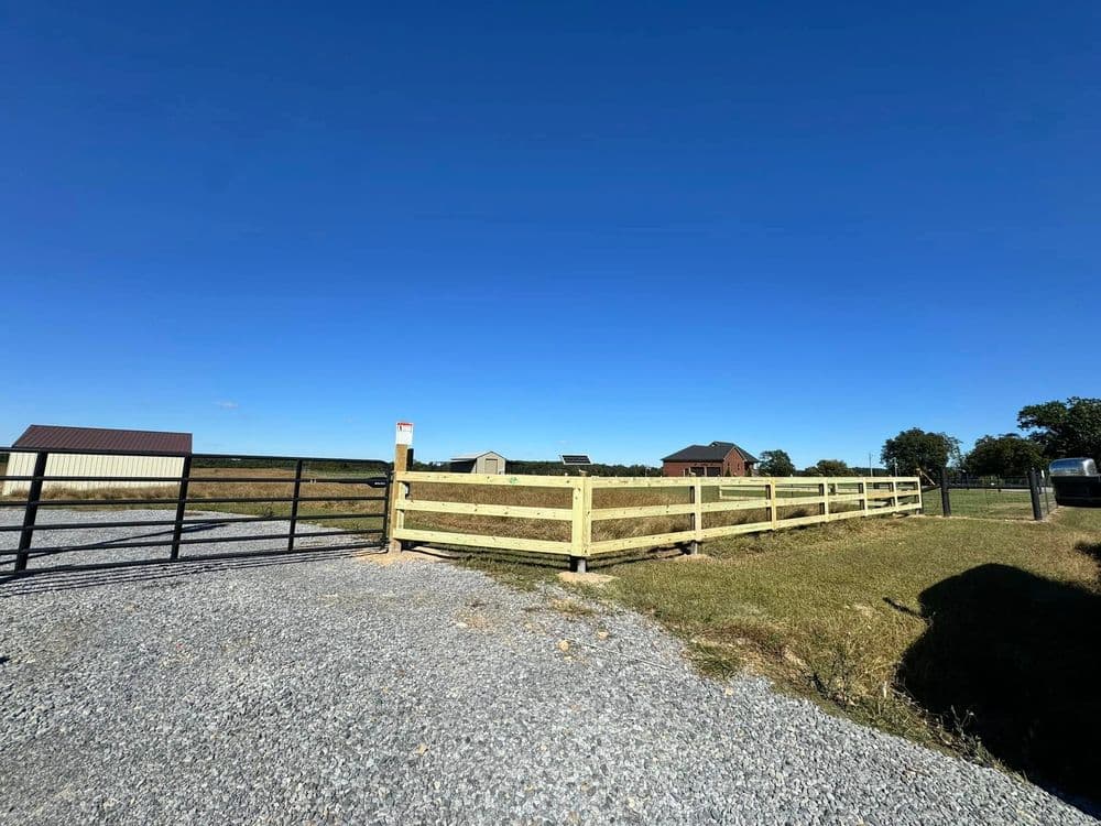 Gravel driveway leading to a wooden fence and open fields under a clear blue sky.