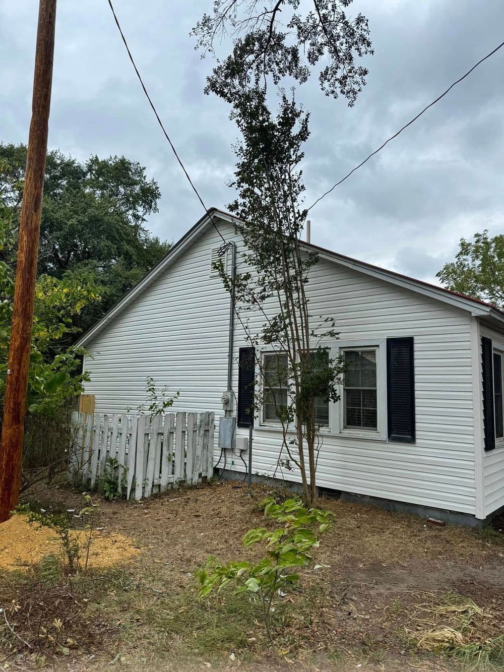 White house with black shutters, surrounded by greenery and utility pole, under cloudy sky.